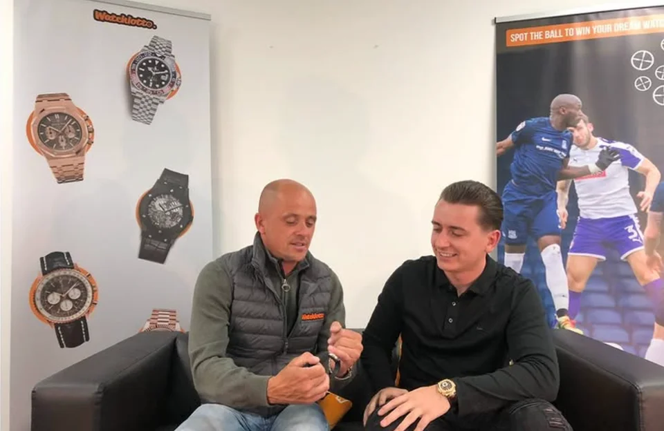 Jamie Robertson and Harrison Gwinnett filming together on a sofa beside Watchlotto banners